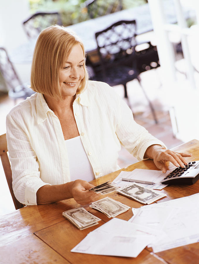 Senior Woman Sitting at a Wooden Kitchen Table, Using a Calculator to Organise Her Personal Finances Photograph by Flying Colours Ltd