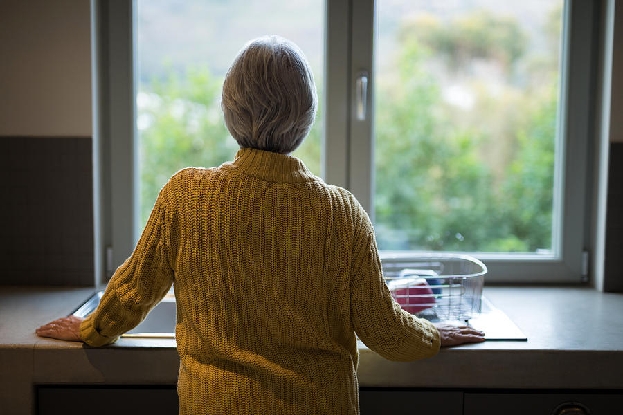 Senior woman standing near the kitchen sink and looking through window Photograph by Wavebreakmedia