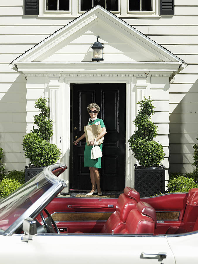 Senior woman standing on porch, convertible car in foreground Photograph by Ryan McVay