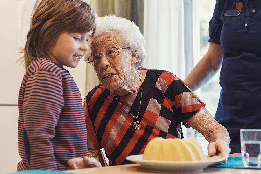 Senior woman talking to great grandson looking at sponge cake on table with caretaker standing at home Photograph by Maskot
