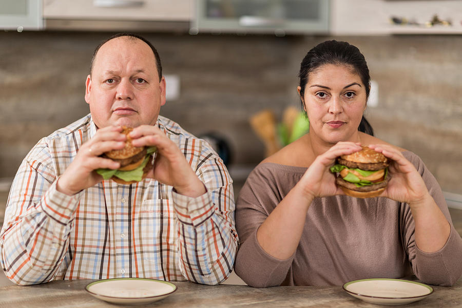 Serious overweight couple eating burgers in the kitchen. Photograph by Skynesher