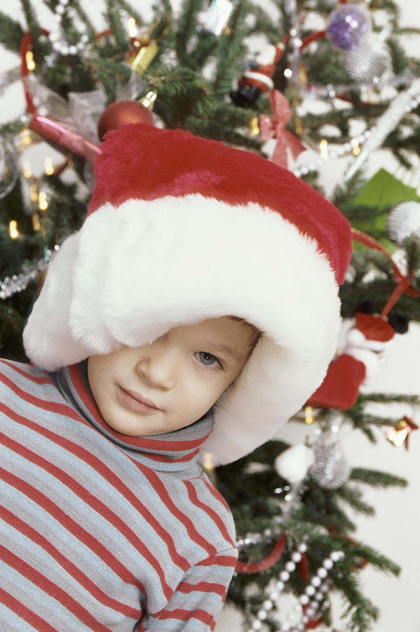 Shy Young Boy Wearing a Big Santa Hat Standing in Front of a Christmas Tree Photograph by B2M Productions
