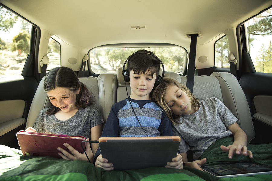 Siblings using digital tablet in back seat of car on road trip Photograph by The Good Brigade