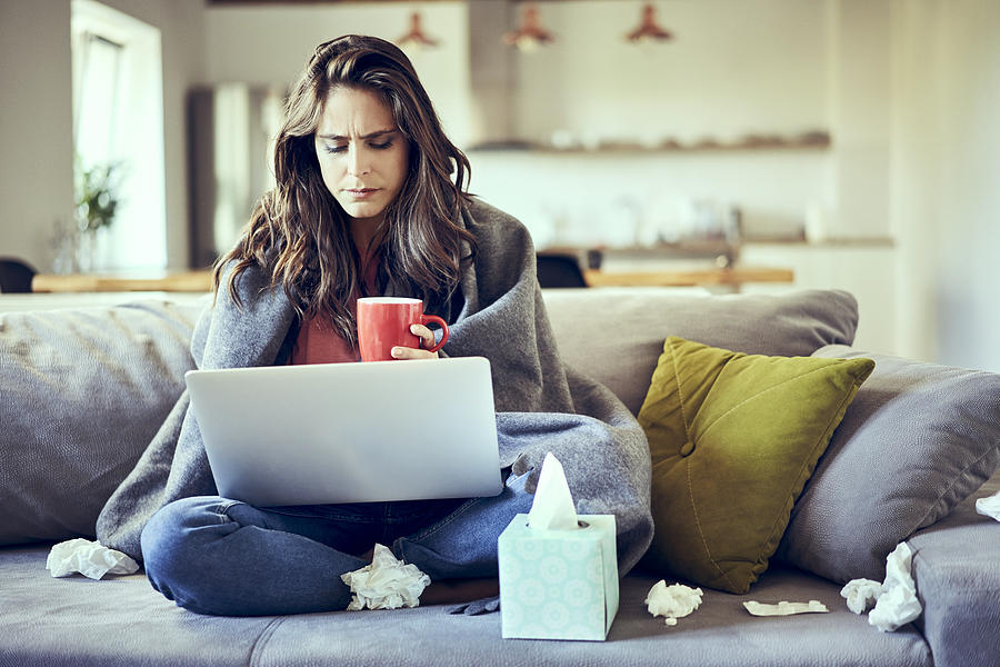 Sick woman sitting on sofa covered in blanket with cup of tea and laptop Photograph by Westend61