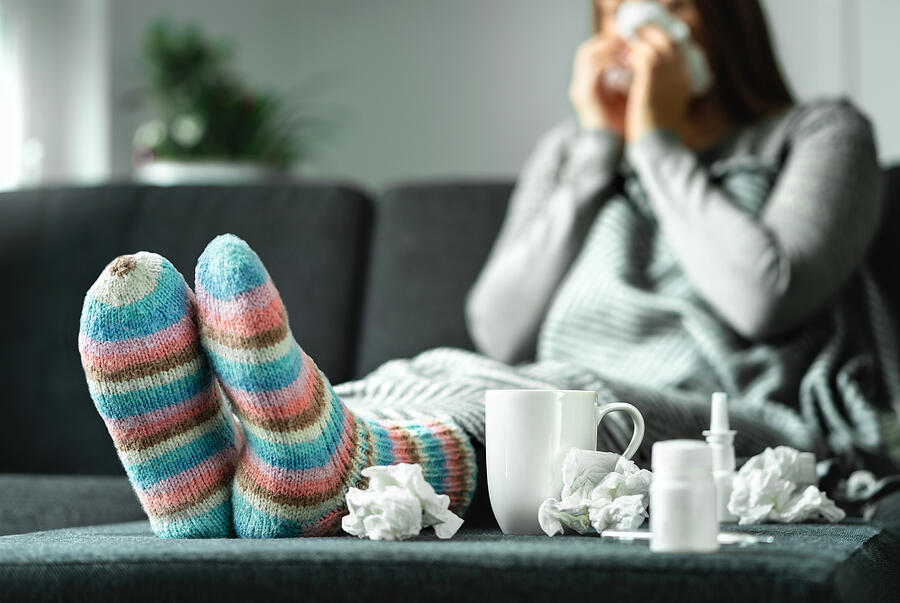 Sick woman with flu, cold, fever and cough sitting on couch at home. Ill person blowing nose and sneezing with tissue and handkerchief. Woolen socks and medicine. Infection in winter. Photograph by Tero Vesalainen
