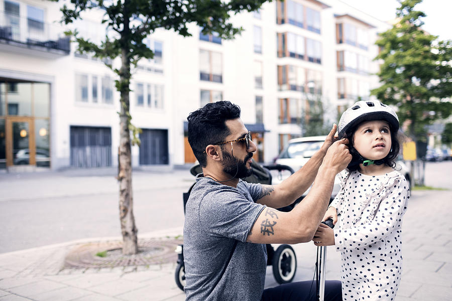 Side view of father assisting daughter in wearing helmet at sidewalk Photograph by Maskot