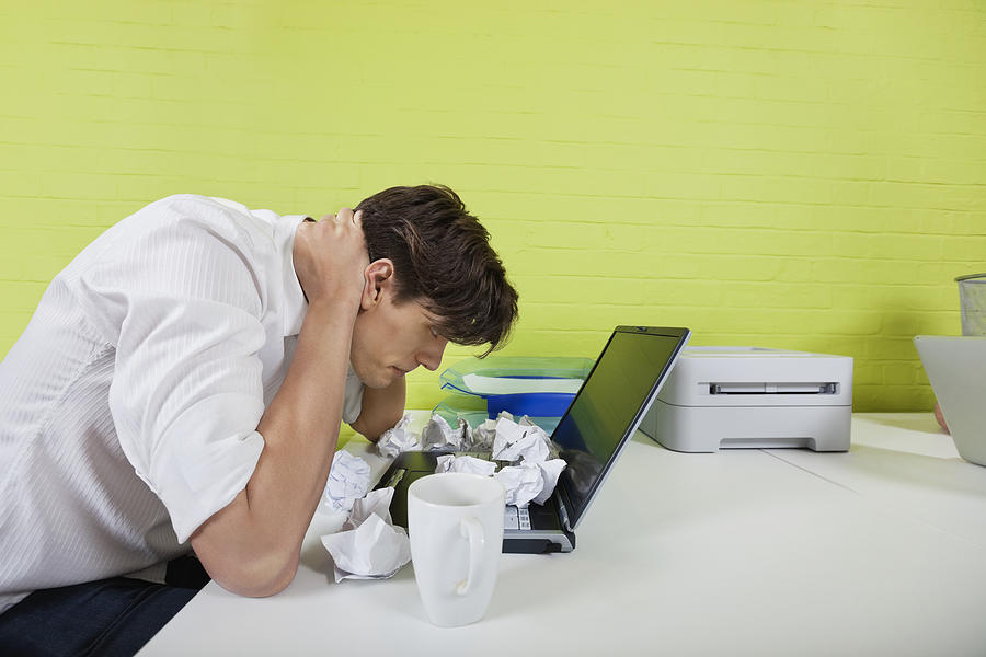 Side view of frustrated young businessman with paper balls and laptop on desk Photograph by Moodboard