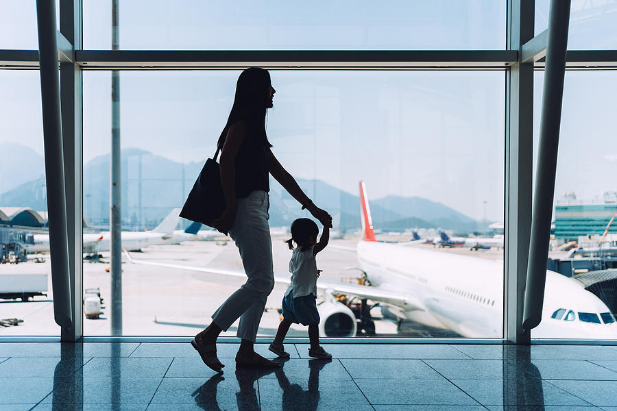 Silhouette of joyful young Asian mother holding hands of cute little daughter looking at airplane through window at the airport while waiting for departure Photograph by D3sign