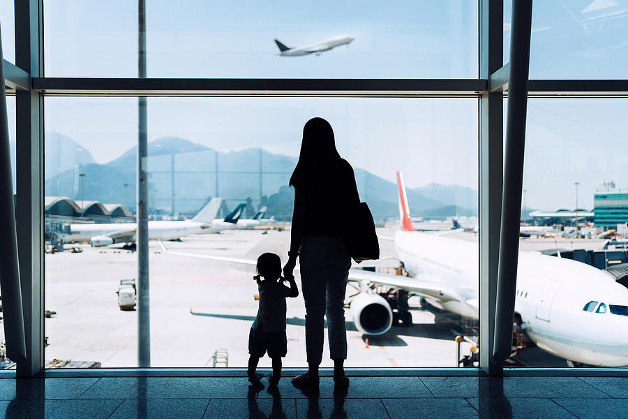 Silhouette of joyful young Asian mother holding hands with cute little daughter looking at airplane through window at the airport while waiting for departure Photograph by D3sign