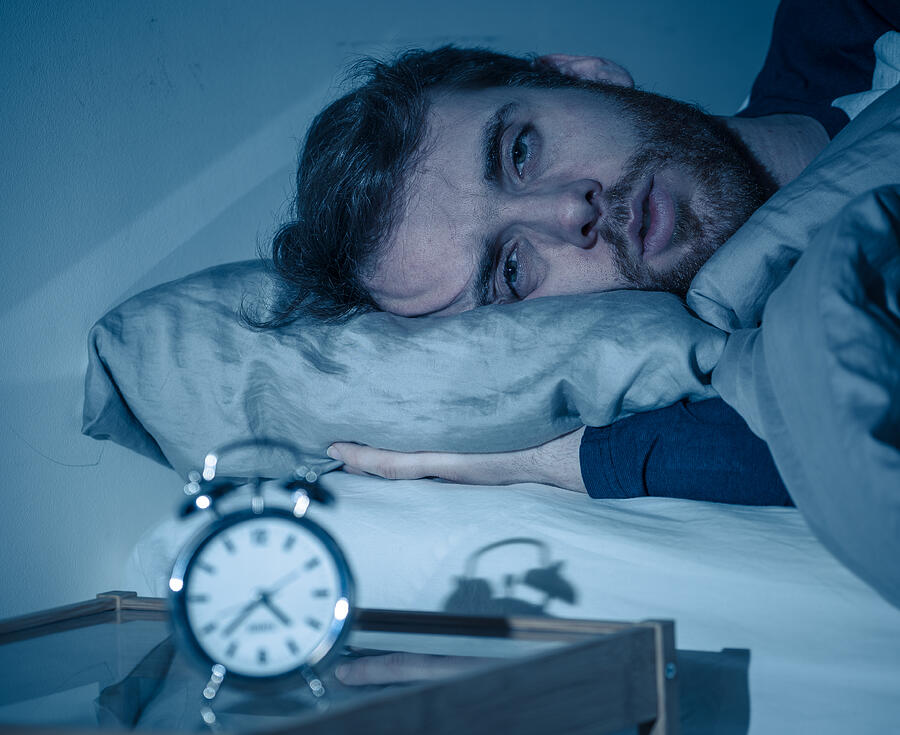 Sleepless and desperate young caucasian man awake at night not able to sleep, feeling frustrated and worried looking at clock suffering from insomnia in stress and sleeping disorder concept. Photograph by SB Arts Media