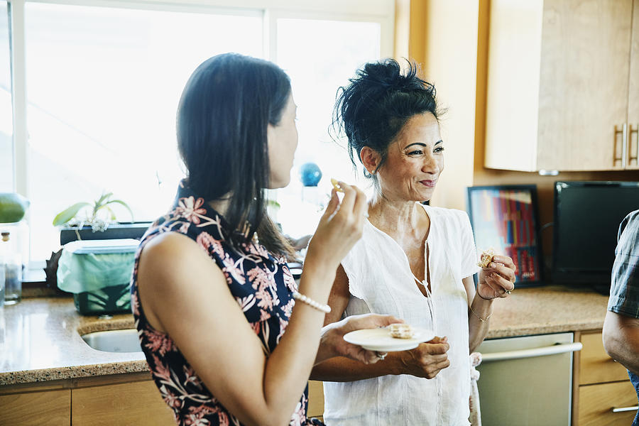 Smiling adult sisters hanging out in kitchen eating lunch Photograph by Thomas Barwick