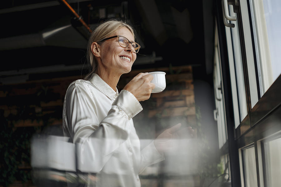 Smiling businesswomanholding cup of coffee looking out of window Photograph by Westend61