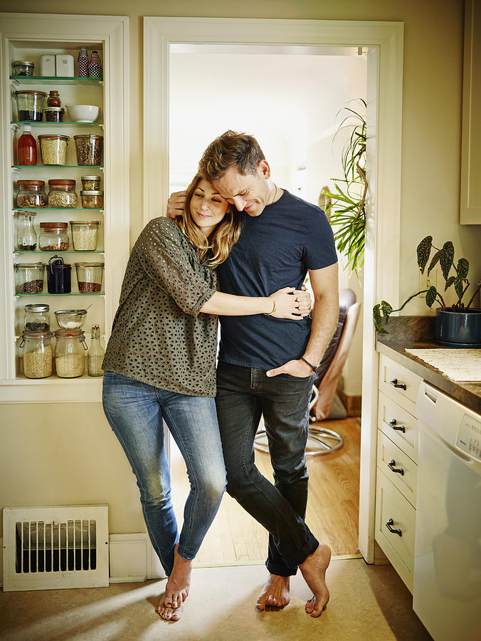 Smiling couple embracing in doorway of kitchen Photograph by Thomas Barwick