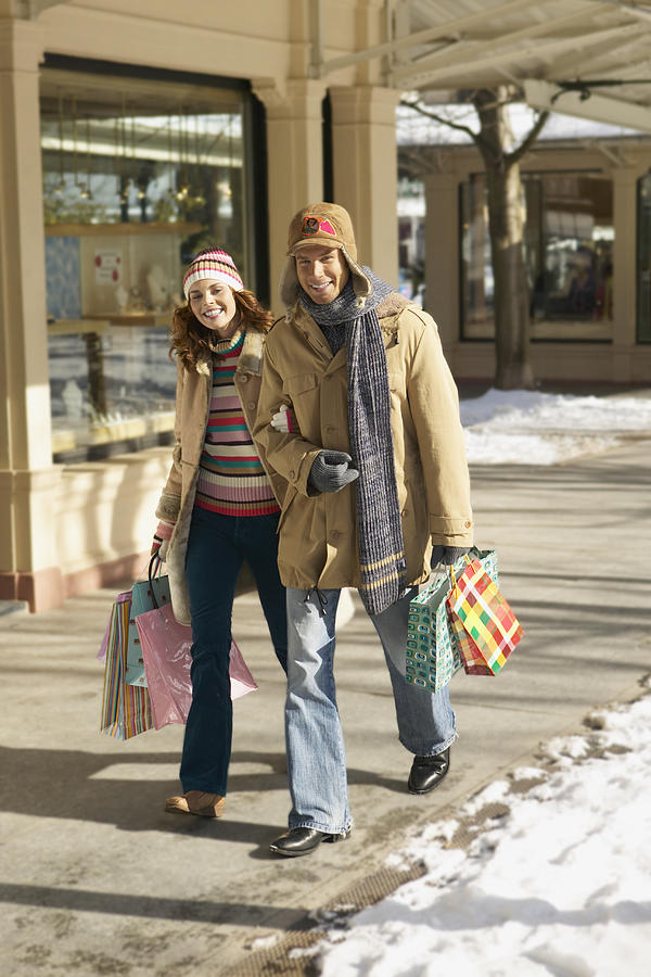 Smiling Couple in Winter Outfits Walk Arm in Arm on the Pavement Carrying Shopping Bags Photograph by Digital Vision.