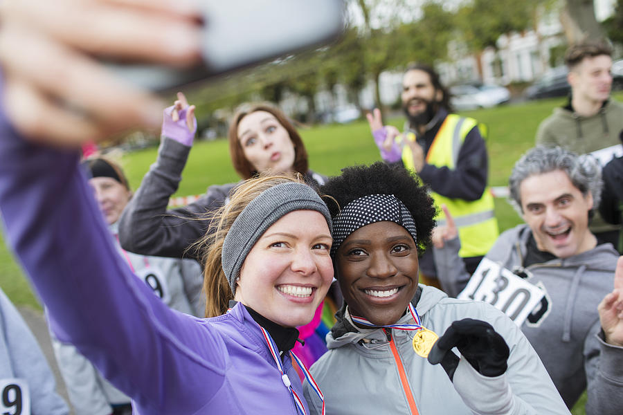 Smiling female runners with medal taking selfie at charity run in park Photograph by Caia Image