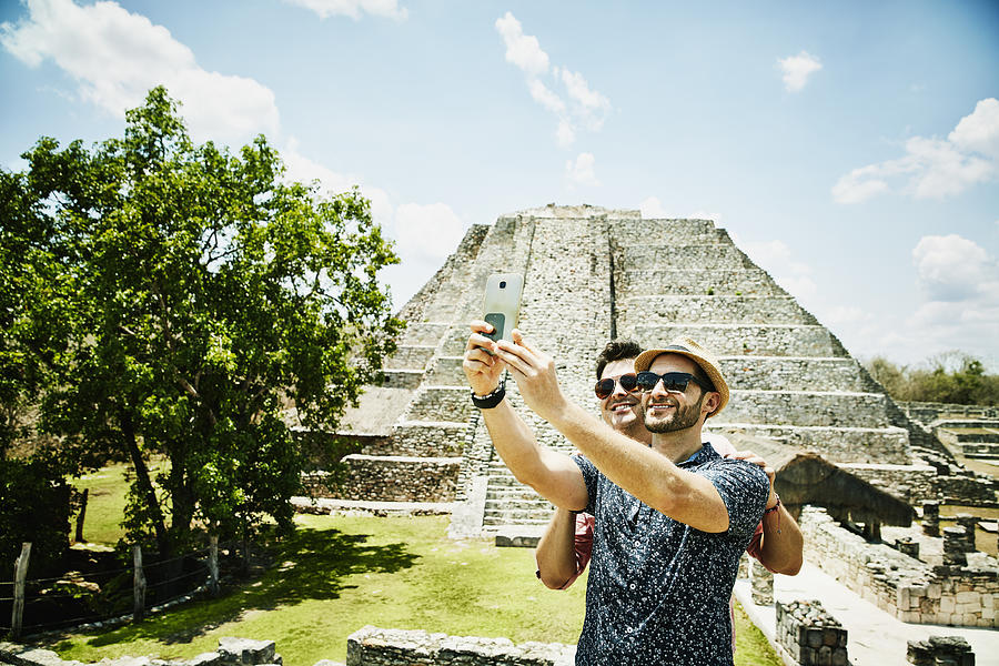 Smiling gay couple taking selfie with smartphone while exploring Mayapan ruins during vacation Photograph by Thomas Barwick
