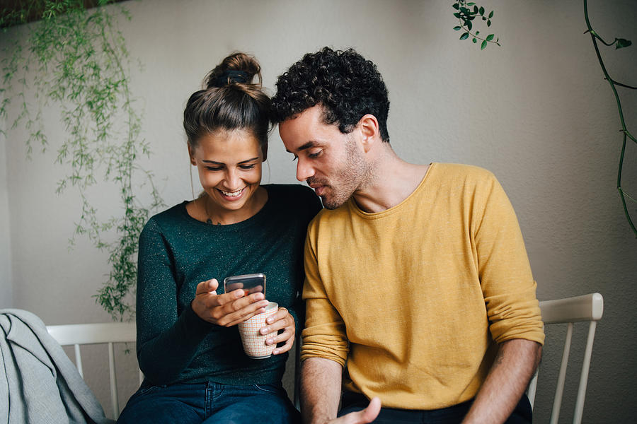 Smiling girlfriend showing smart phone to boyfriend while having coffee in living room Photograph by Maskot