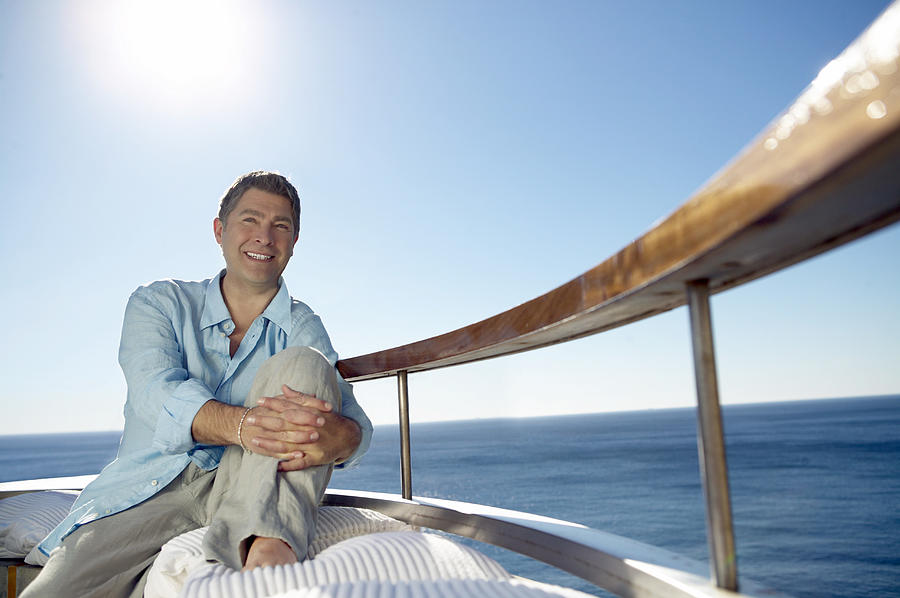 Smiling Man in Casual Summer Clothing Sits on a Sunlit Balcony by the Sea Photograph by Digital Vision.