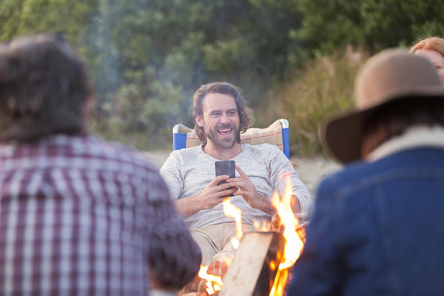 Smiling man using cell phone on beach by camp fire Photograph by Compassionate Eye Foundation/Steven Errico