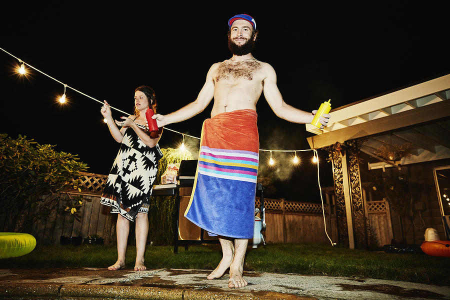 Smiling man wrapped in towel holding ketchup and mustard while grilling for friends in backyard during party on summer evening Photograph by Thomas Barwick