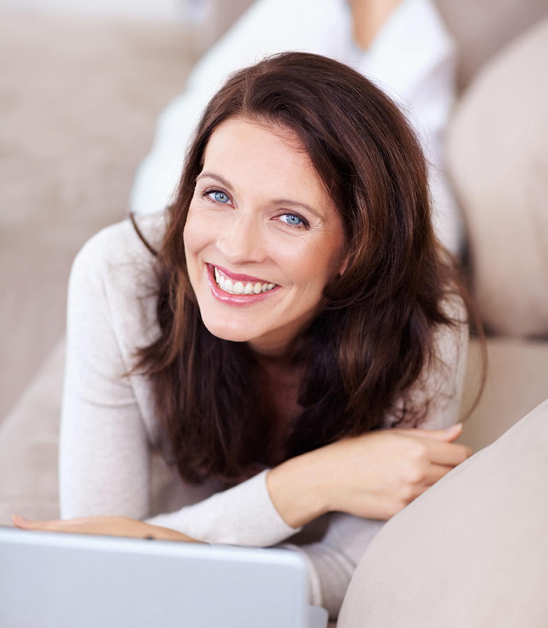 Smiling mature lady lying on couch and using laptop Photograph by Jacob Wackerhausen