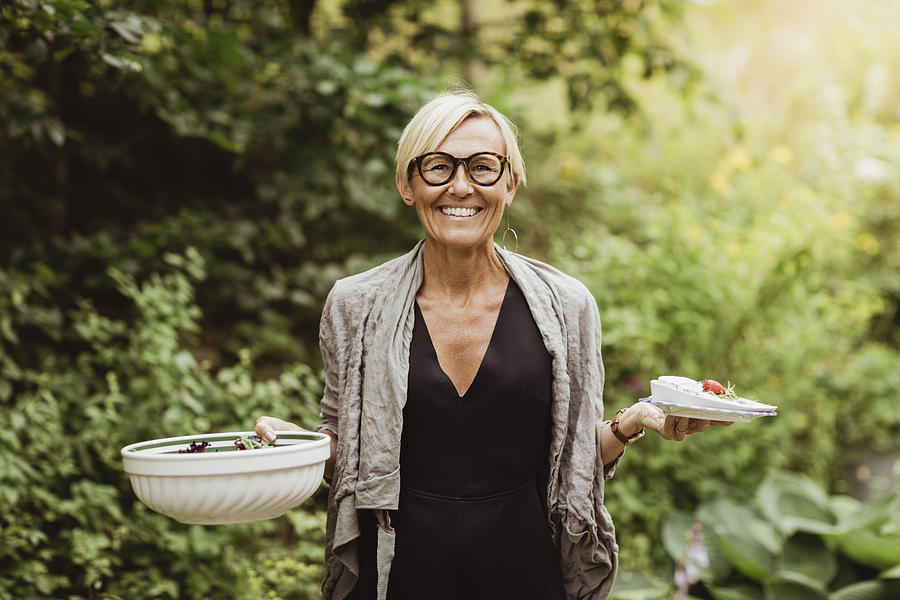 Smiling mature woman holding food in bowl and plate against plants in front yard Photograph by Maskot
