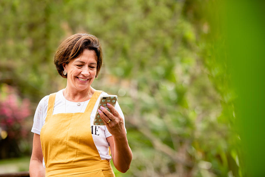 Smiling mature woman video calling on a phone outside in her yard Photograph by Giselleflissak