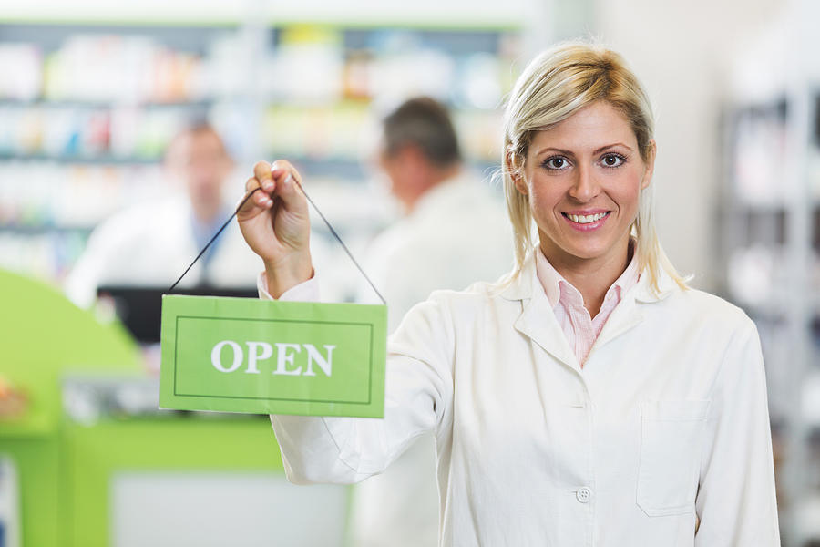 Smiling pharmacist holding open sign in pharmacy. Photograph by Skynesher