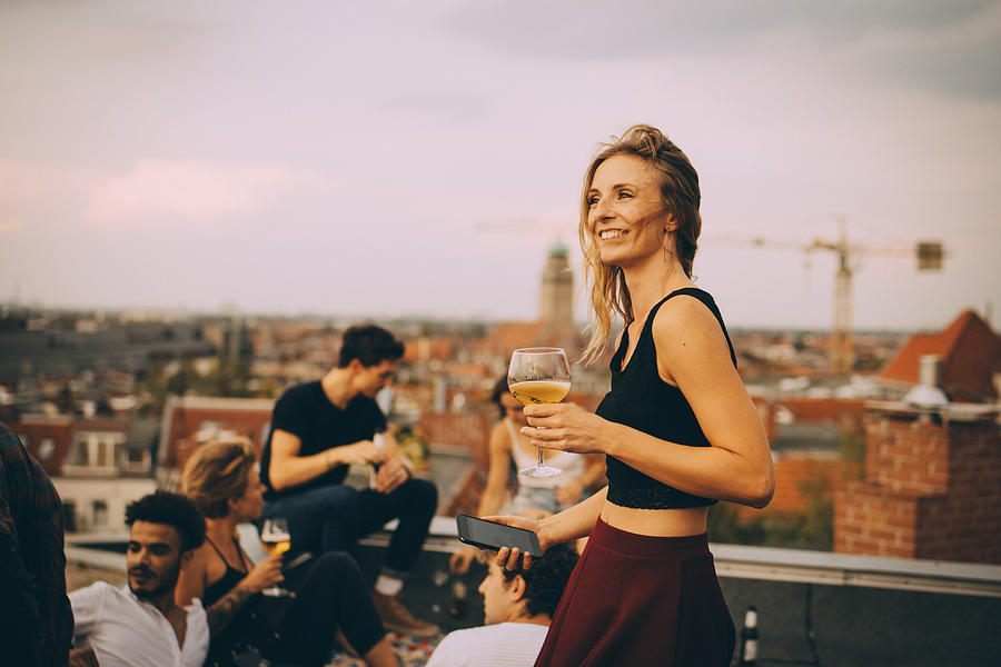 Smiling woman enjoying drink while partying with friends at rooftop Photograph by Maskot