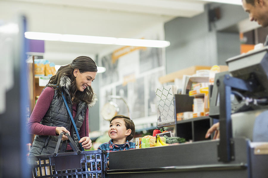 Smiling woman holding basket while standing with son by checkout counter at supermarket Photograph by Cavan Images