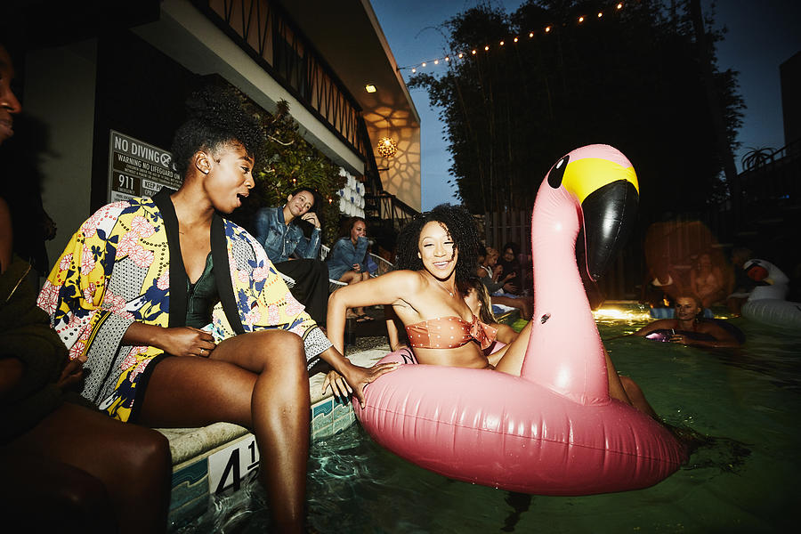Smiling woman sitting in inflatable flamingo in pool during party with friends at hotel Photograph by Thomas Barwick