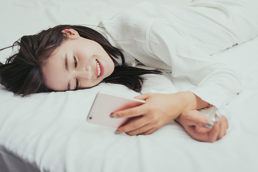 Smiling woman texting on cell phone on bed Photograph by Xuanyu Han