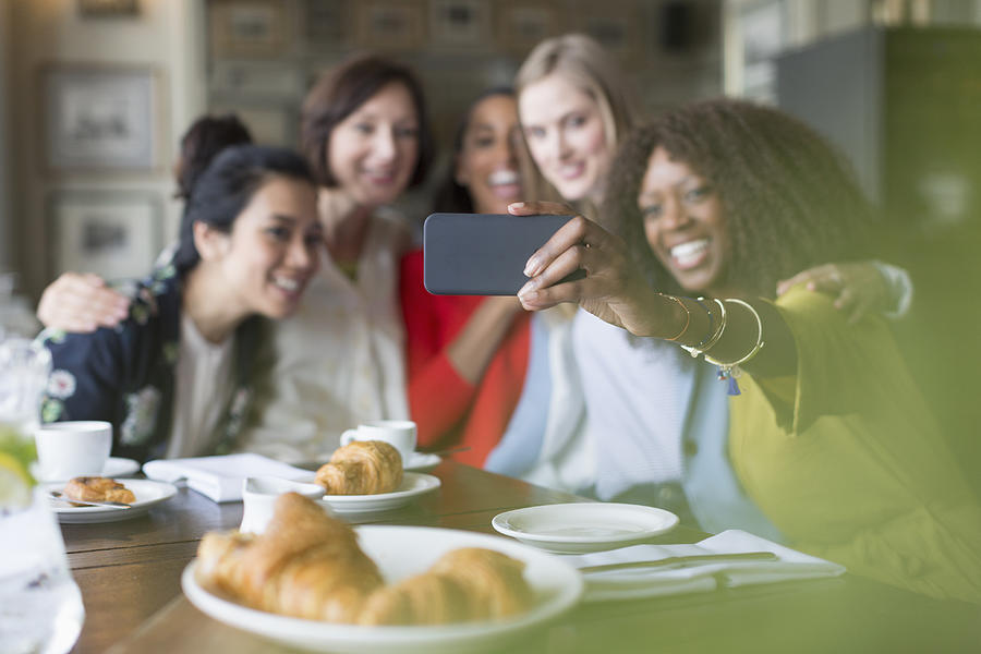 Smiling women friends taking selfie with camera phone in restaurant Photograph by Caia Image