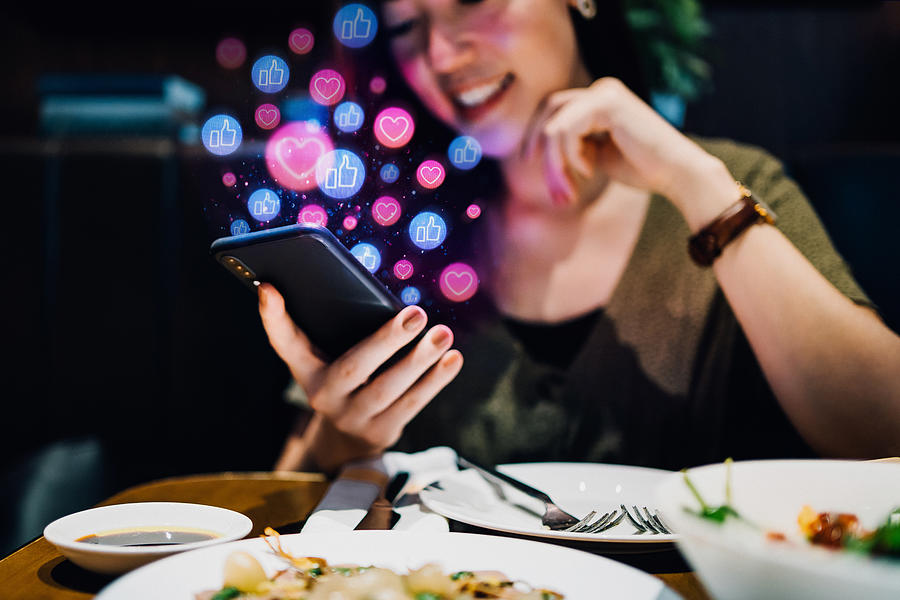 Smiling young Asian woman using smartphone on social media network application while having meal in the restaurant, viewing or giving likes, love, comment, friends and pages. Social media addiction concept Photograph by D3sign