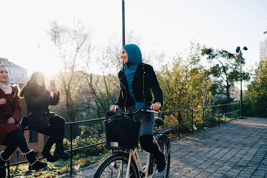 Smiling young woman cycling on footpath by female friends sitting on railing in city Photograph by Maskot