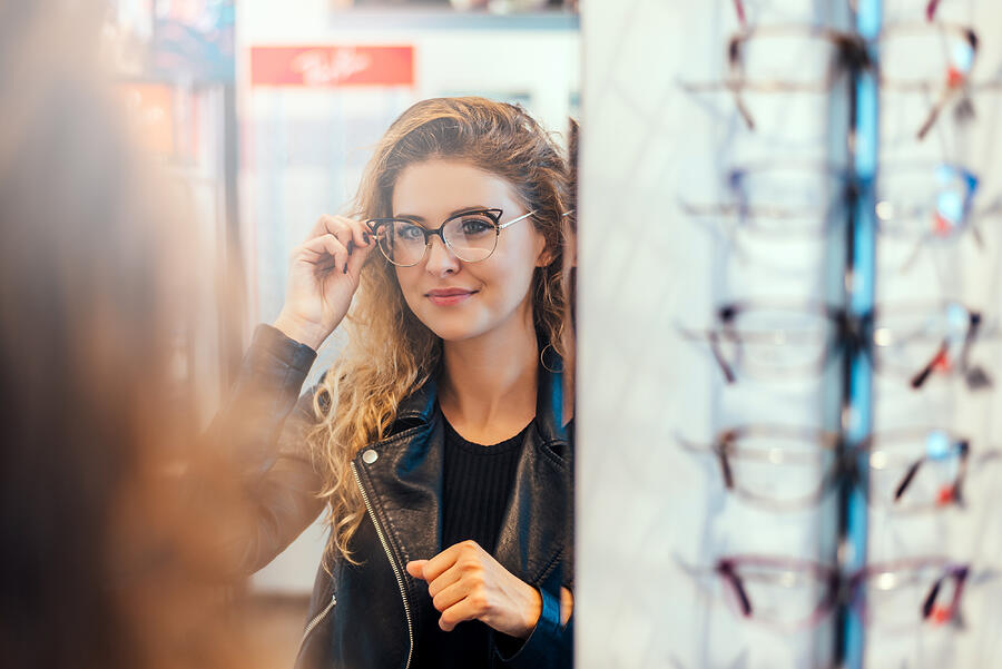 Smiling young woman trying on glasses on mirror in optician. Photograph by Nortonrsx