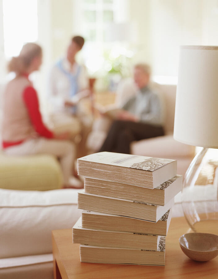 Stack of books sitting on table in living room, women in background Photograph by Victoria Pearson
