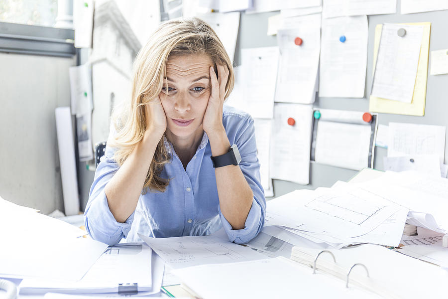 Stressed woman sitting at desk in office surrounded by paperwork Photograph by Westend61