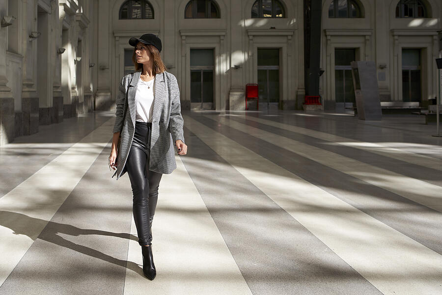 Stylish woman wearing white t-shirt, long blazer, black leather pants and black cap walking alone through a public corridor. Photograph by VEAM Visuals