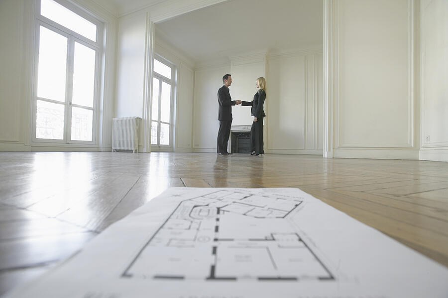 Surface Level Shot of Blueprints on a Wooden Floor in an Empty Room, with a Businessman Shaking Hands with a Businesswoman in the Background Photograph by B2M Productions