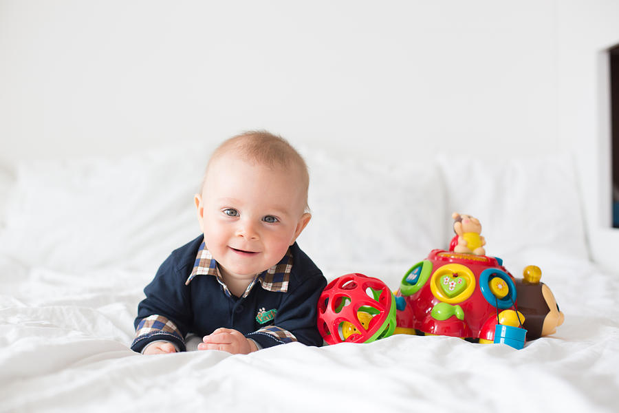 Sweet toddler boy, playing with colorful toys in bed, daytime. Childhood happiness concept, early development toys Photograph by Tatyana Tomsickova Photography