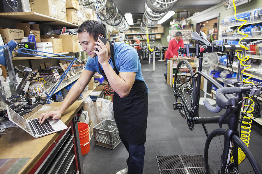 Technician using cell phone and laptop in bicycle repair shop Photograph by Jetta Productions Inc