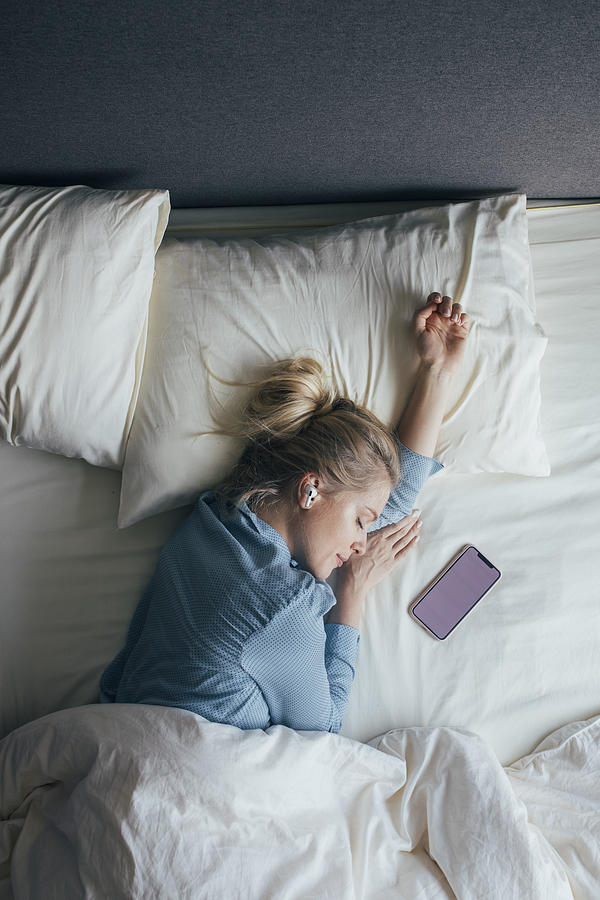 Technology-Aided Sleep: Woman in Pyjamas With Wireless Headphones Listening to Relaxing Music on Her Smartphone in Bed in the Morning Photograph by FreshSplash