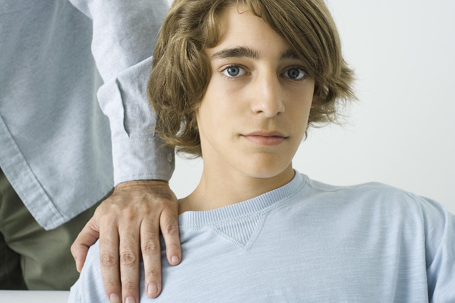 Teen boy with mans hand on his shoulder, looking at camera Photograph by PhotoAlto/Frederic Cirou