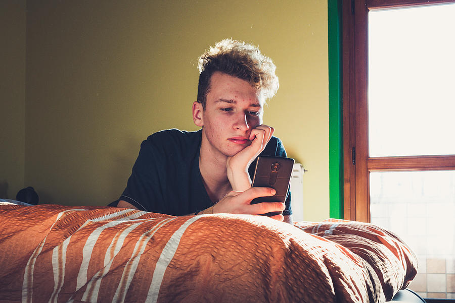 Teenage Boy Using his Smart Phone while Laying on the Bed Photograph by CasarsaGuru