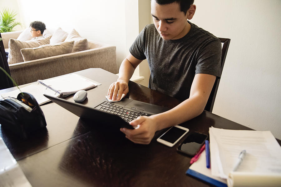 Teenage boy wiping laptop while studying from home. Photograph by Martinedoucet