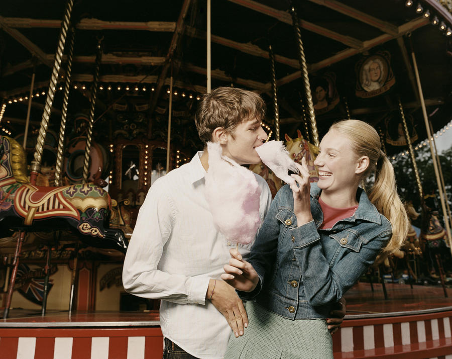 Teenage Couple Stand in Front of a Carousel, Girl Feeding Her Boyfriend Candy Floss and Laughing Photograph by Digital Vision.