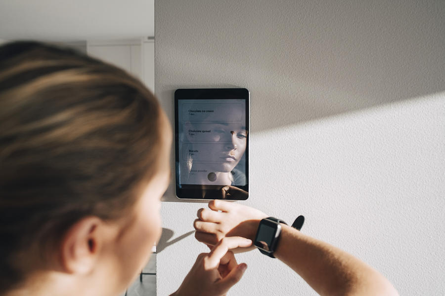 Teenage girl adjusting smart watch with digital tablet mounted on white wall at home Photograph by Maskot