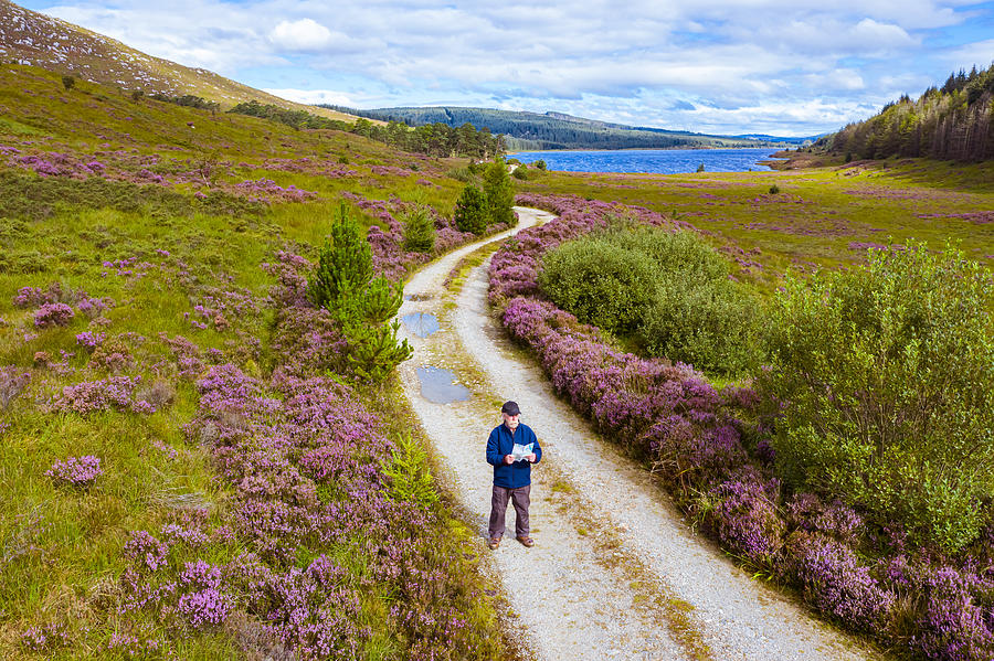 The high angle view of an active senior man looking at a map while standing on a dirt road in a remote part of Dumfries and Galloway, south west Scotland Photograph by JohnFScott