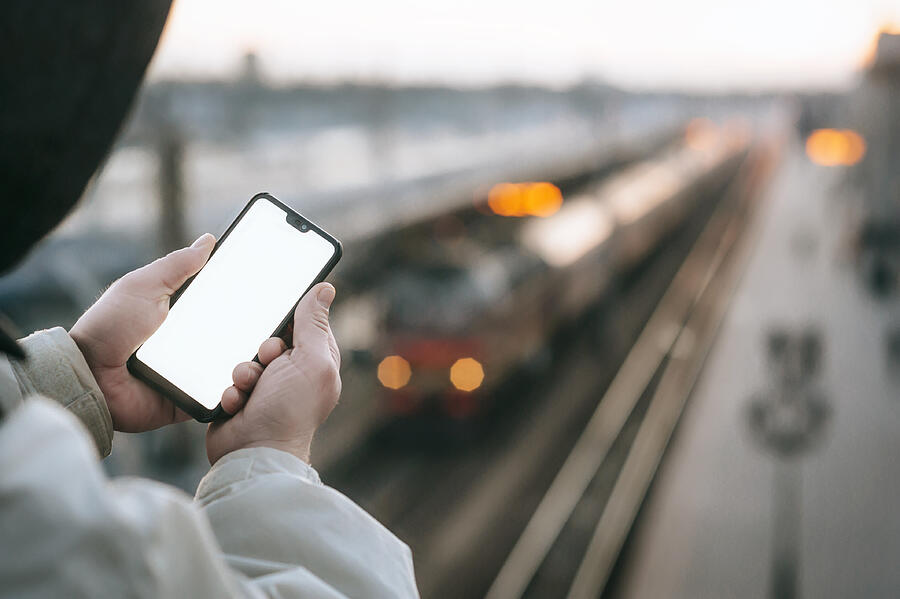 The Man is holding a mock up smartphone in his hand, against the background of the train at the railway station. Photograph by Victor Prilepa
